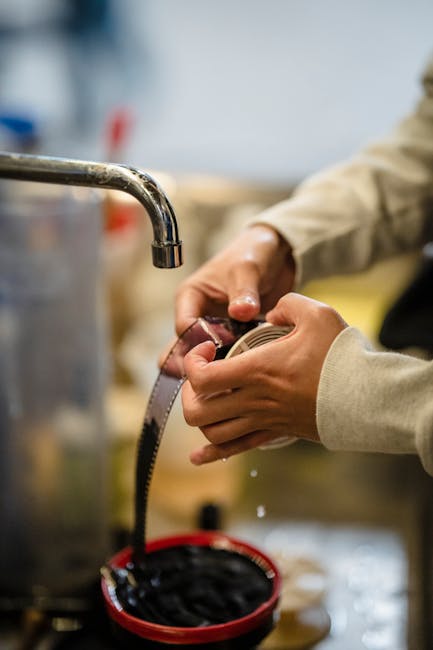 A person wearing a beige long-sleeve shirt is holding a coiled measuring tape, preparing to measure a piece of furniture or packaging material in a home environment. The scene is set indoors, with a metal faucet visible on the left side of the image, suggesting proximity to a sink or kitchen area. The background is blurred but includes various cardboard boxes and packing supplies, indicating an ongoing packing or moving process as part of home relocation or furniture transport. The lighting is natural or well-lit, highlighting the hands and the tools used in the packing or loading process. This visual relates to professional removals services provided by Removals Ilford, focused on careful packing, measuring, and preparing items for safe transit during house moves, in line with the topic of avoiding hidden removal fees in Ilford.