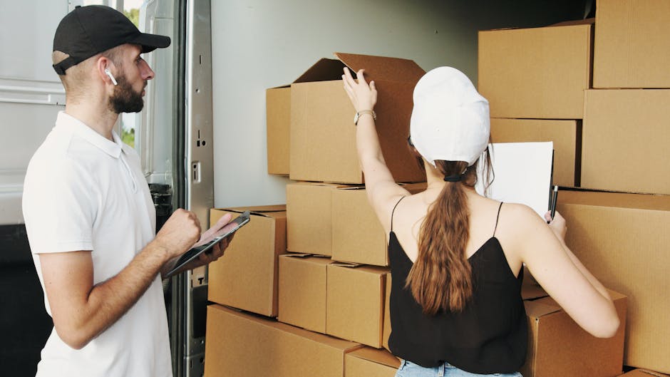 A professional moving team preparing for a home relocation process outdoors, with two individuals inspecting and organizing cardboard moving boxes stacked on a pavement near a van. The man, wearing a black cap, white polo shirt, and earbuds, holds a clipboard and appears to be checking inventory or planning the loading process. The woman, with long hair tied back and wearing a white protective hat and black sleeveless top, is reaching up to adjust or check a large cardboard box. The scene includes a large metal van with an open sliding door, revealing part of the vehicle's interior used for furniture and box transport, with the environment being well-lit by natural daylight. This image demonstrates the packing, inventory management, and loading activities involved in furniture transport and house removals, as conducted by [COMPANY_NAME], highlighting the logistics of careful box handling and vehicle loading typical in professional removal services in Ilford.