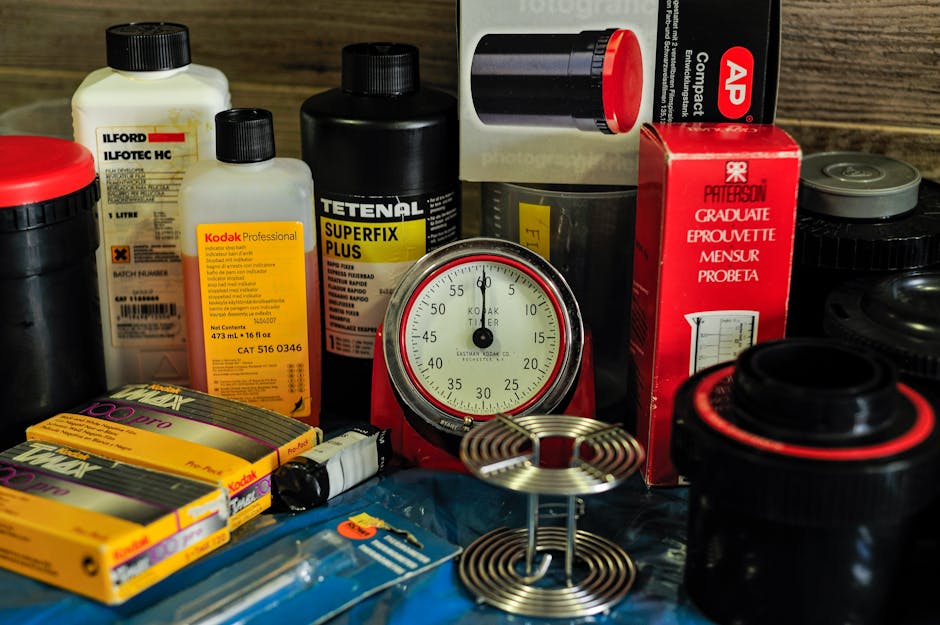A collection of laboratory and photographic chemicals, supplies, and equipment displayed on a flat surface, including bottles of Ilford photographic chemicals, Kodak Professional film, and Tetenal Superfix Plus liquid, situated in an indoor environment. The bottles are made of plastic with black caps, some labels detailed with product information. A round, red-and-white mechanical timer is centrally positioned among the items. Several plastic and cardboard packaging are visible, with some boxes partially open or stacked. The scene is well-lit, highlighting the various textures and materials such as glass, plastic, and cardboard. This arrangement of supplies and equipment suggests a setting related to image processing or scientific work, which may be relevant in the context of packing or transferring similar household or professional items during a home relocation, as handled by [COMPANY_NAME] specializing in removals and moving services. Located on a wooden surface, the focus is on accurately describing the objects and their environment for accessibility and relevance to house removals and furniture transport processes.