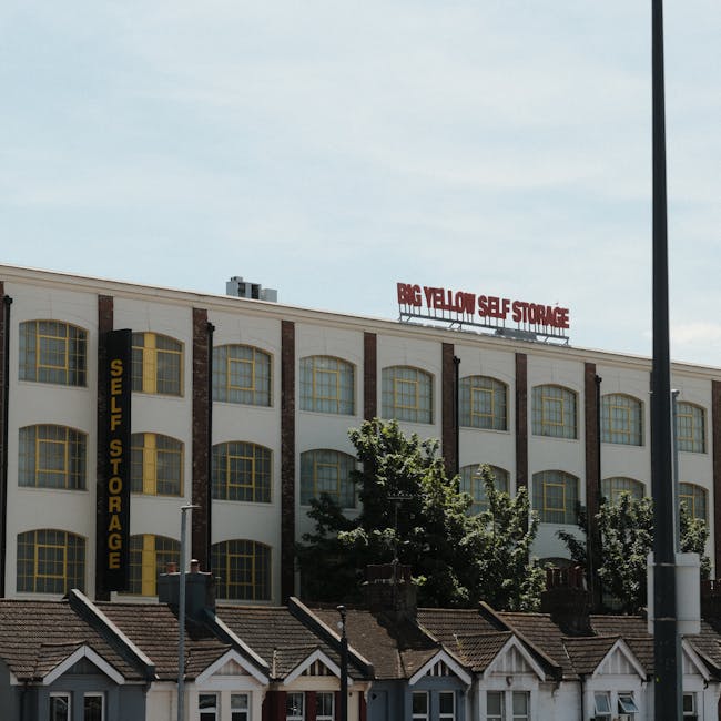 Photograph of a large three-story building hosting a self-storage facility in Ilford, with a white facade accented by brick vertical stripes and several large yellow-framed arched windows on each level. Prominent signage for 'Big Yellow Self Storage' is visible at the top of the building in red letters, while a vertical yellow sign on the side reads 'Self Storage.' Below, a row of small residential houses with tiled roofs and chimneys line the street at the building's base. The scene shows clear daylight with a partly cloudy sky. Trees partially obscure the lower portion of the storage facility, and a tall black street lamp and a pole are seen in the foreground, suggesting the location is within an urban area. This image relates to house removals and furniture transport services, reflecting the context of relocation and moving logistics, with the visual emphasis on storage solutions often used during home relocations.