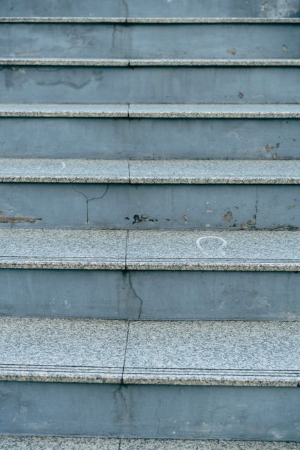 A close-up view of a weathered concrete staircase outdoors, with six visible steps featuring chipped edges, cracks, and signs of wear. The steps are painted in a faded blue-gray color, with some areas