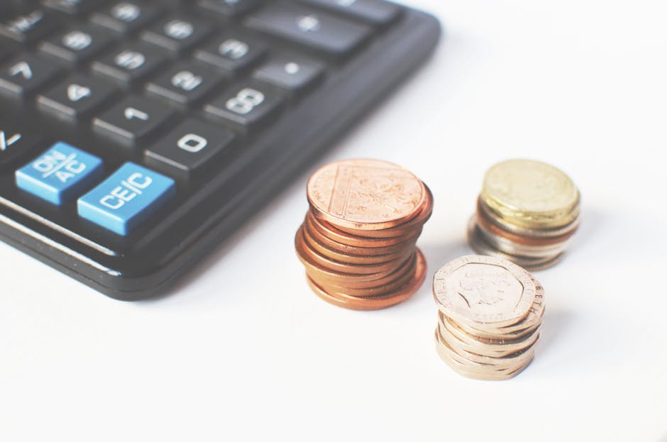 A close-up image showing a black calculator with blue and white buttons on the left side, positioned on a white surface. To the right of the calculator, there are multiple stacks of coins, including c