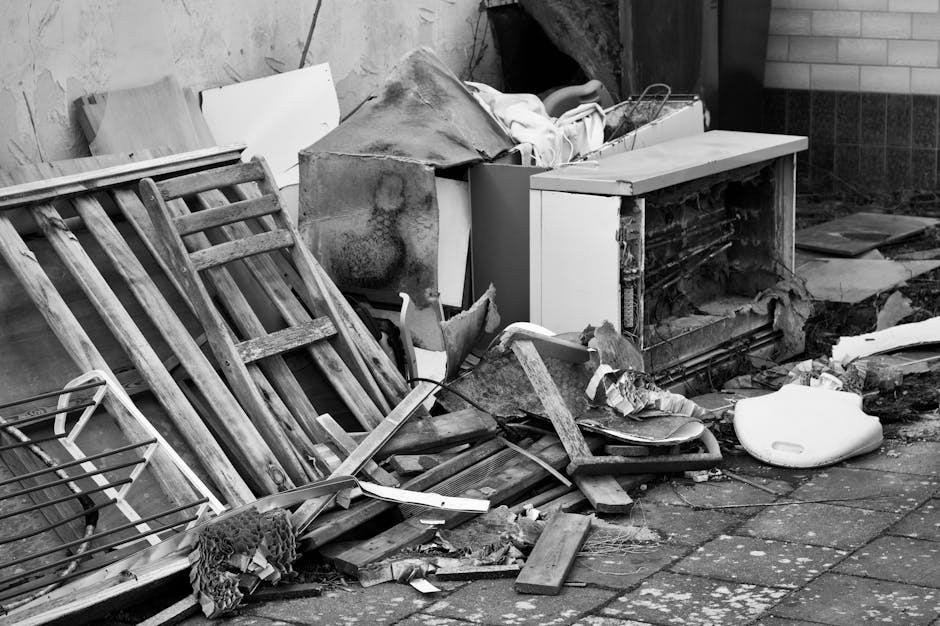 A cluttered outdoor area next to a building during the loading process for house removal services, with various discarded household items and building debris. Visible objects include a disassembled wooden bed frame leaning against a wall, a damaged cabinet with its door missing, and a pile of cardboard boxes, some torn or flattened, alongside plastic wrapping and packaging materials. The ground is a paved surface with scattered debris and loose wood pieces, indicating ongoing clearance or packing work. In the background, part of a brick wall and a section of the pavement are visible under natural lighting. This image exemplifies the type of gathered waste or unwanted items handled during a home relocation or furniture transport process, as managed by removals companies like Removals Ilford, in accordance with local waste disposal rules and moving logistics for property clearance.