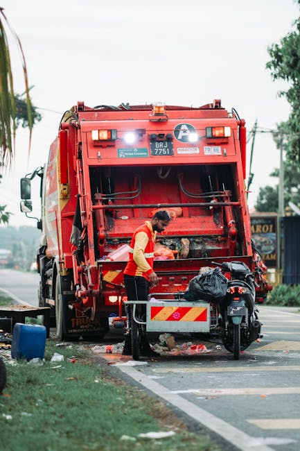 A sanitation worker in a high-visibility red and yellow uniform is standing behind a large red refuse collection vehicle on a paved road, near a grassy verge. The worker is using their hands to operate the rear loading mechanism of the trash truck, which is open and emptying its contents. Nearby, on the pavement, there is a black plastic trash bag and some scattered debris. The vehicle is equipped with various safety lights and reflective strips, with the registration plate 'BRJ 7751' prominently displayed at the back. In the background, there are trees, utility poles, and a few buildings, indicating a suburban or urban residential area. The scene occurs during daytime, with overcast weather and natural lighting. This image reflects home waste disposal and the process of rubbish collection, relevant to house removals and waste management services provided by Removals Ilford, highlighting proper waste handling during property relocations as per local regulations.