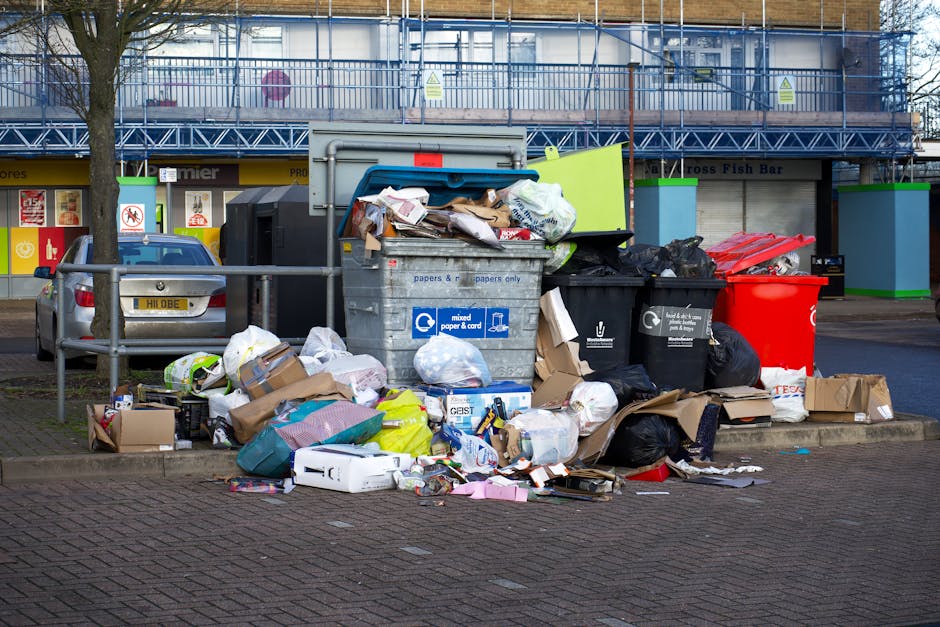 A cluttered outdoor area next to a building during the loading process for house removal services, with various discarded household items and building debris. Visible objects include a disassembled wooden bed frame leaning against a wall, a damaged cabinet with its door missing, and a pile of cardboard boxes, some torn or flattened, alongside plastic wrapping and packaging materials. The ground is a paved surface with scattered debris and loose wood pieces, indicating ongoing clearance or packing work. In the background, part of a brick wall and a section of the pavement are visible under natural lighting. This image exemplifies the type of gathered waste or unwanted items handled during a home relocation or furniture transport process, as managed by removals companies like Removals Ilford, in accordance with local waste disposal rules and moving logistics for property clearance.