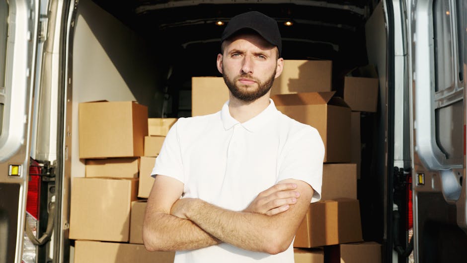 A young man with a beard wearing a white polo shirt and black cap stands inside a large moving van with its rear doors open, revealing stacked cardboard boxes of various sizes secured and organized for a house removal. He is crossing his arms and looking directly at the camera with a serious expression. The interior of the van is illuminated with natural light, showing a neat arrangement of packing materials and boxes prepared for home relocation. The scene reflects the process of furniture transport and packing and moving services provided by Removals Ilford, with the van positioned on a paved driveway or street adjacent to the property. This image illustrates the logistical aspects of a tight access move, highlighting the importance of careful handling and organized loading during furniture transport and home relocation projects.