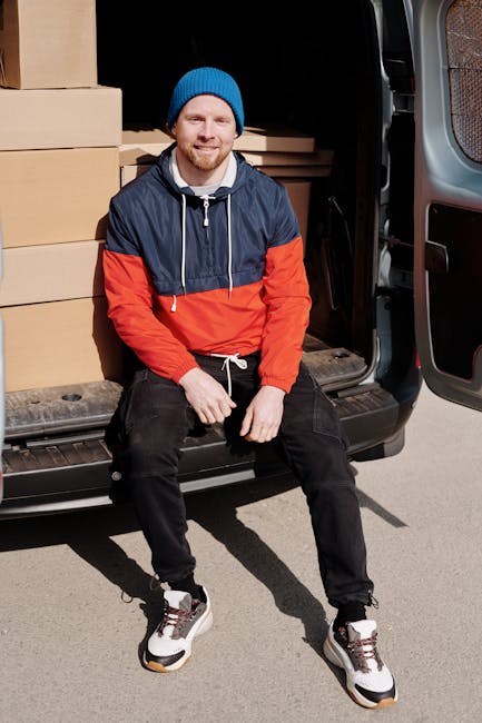 A young man with a beard wearing a white polo shirt and black cap stands inside a large moving van with its rear doors open, revealing stacked cardboard boxes of various sizes secured and organized for a house removal. He is crossing his arms and looking directly at the camera with a serious expression. The interior of the van is illuminated with natural light, showing a neat arrangement of packing materials and boxes prepared for home relocation. The scene reflects the process of furniture transport and packing and moving services provided by Removals Ilford, with the van positioned on a paved driveway or street adjacent to the property. This image illustrates the logistical aspects of a tight access move, highlighting the importance of careful handling and organized loading during furniture transport and home relocation projects.