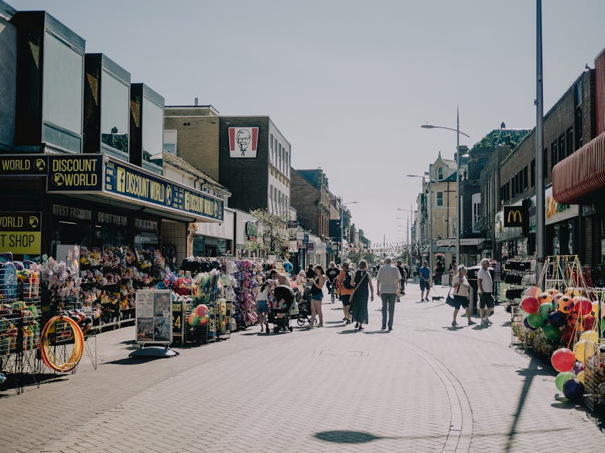 A busy street scene outside a retail area with several small shops and stalls, including a discount store and a McDonald's. The storefronts are displaying various merchandise, such as balloons, toys, and flowers, arranged on tables and racks along the pavement. People are walking along the street, some pushing strollers, others carrying shopping bags, and a few are browsing the outdoor displays. The street is lined with modern and older style buildings, with signage visible for different brands and shops. On the right side, there is a lamppost and part of a closed storefront with a striped awning. The image showcases a typical urban shopping district during daylight, with clear, bright conditions highlighting the scene's activity related to local shopping, which could be part of furniture or equipment moving activities by Removals Ilford working to facilitate home or business relocations nearby.