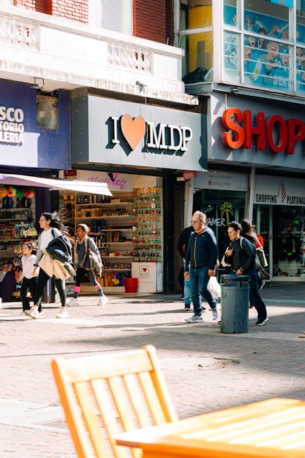Street scene in Ilford showing pedestrians walking past retail shops with signage for 'I ♥ MDP' and other storefronts, including a hardware or gift shop. The storefronts feature large windows displaying various products and bright signage, with some window displays visible. In the foreground, part of an outdoor seating area with a yellow wooden table and chair is visible, slightly out of focus. The sidewalk is lined with a trash bin and a nearby clothing or accessory store can be seen. The scene depicts a busy commercial area during daytime, with individuals of different ages and styles of dress, some carrying shopping bags or backpacks, walking in multiple directions. The overall setting is urban with natural daylight illuminating the scene, typical of an active shopping district in Ilford. This context relates to local commercial activity and reflects the type of environment where house removals or furniture transport services by Removals Ilford might operate.